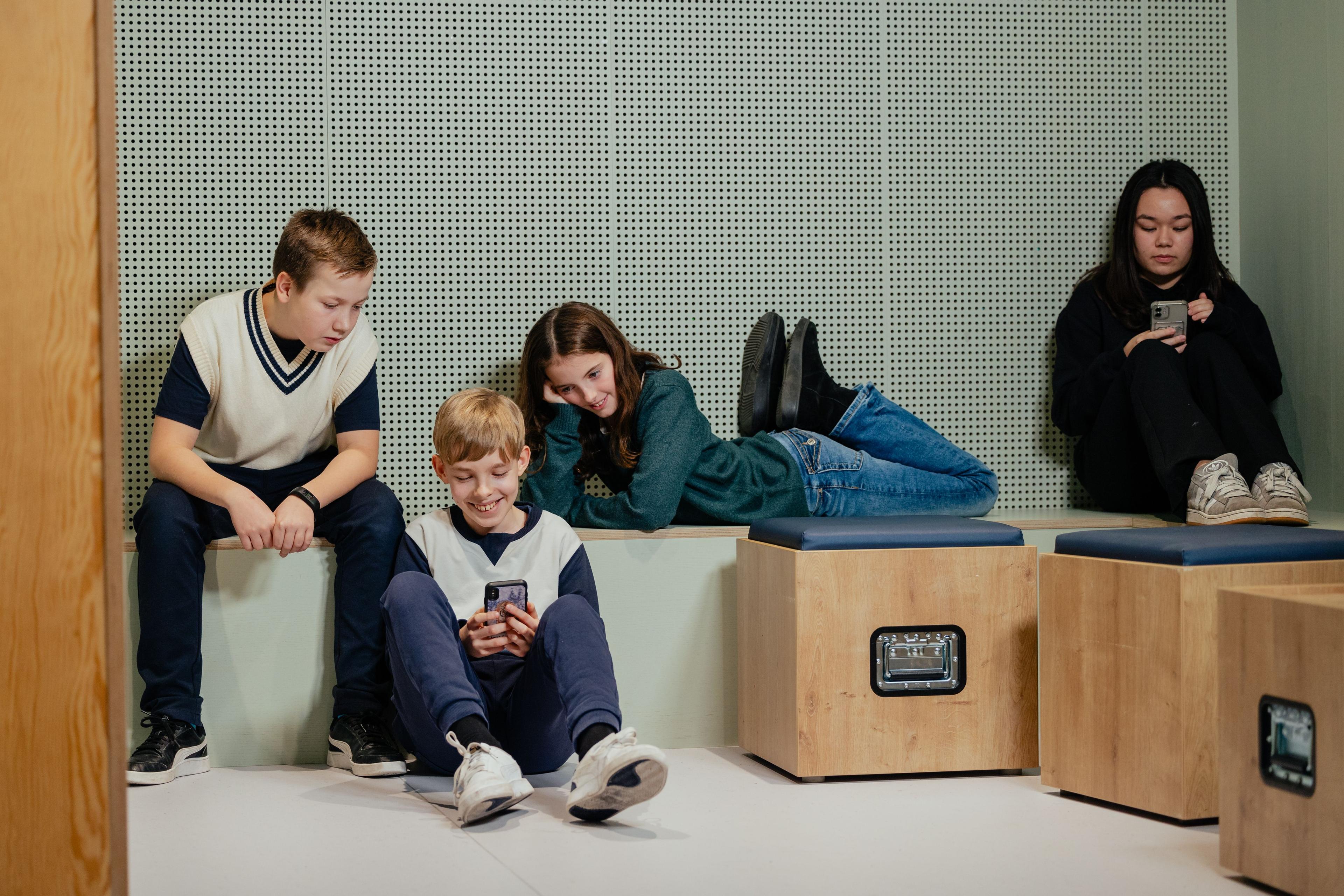 Tre børn sidder sammen og ser på en telefon imens en anden pige slapper af på sin egen telefon